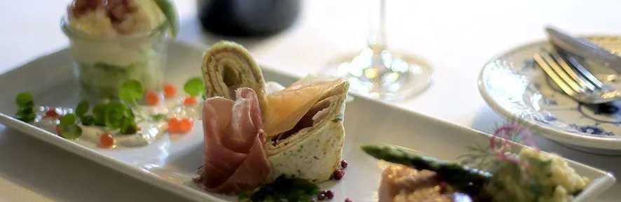 En elegant tallerken med gourmetretter, herunder skinke, asparges og en lækker dessert i glas.