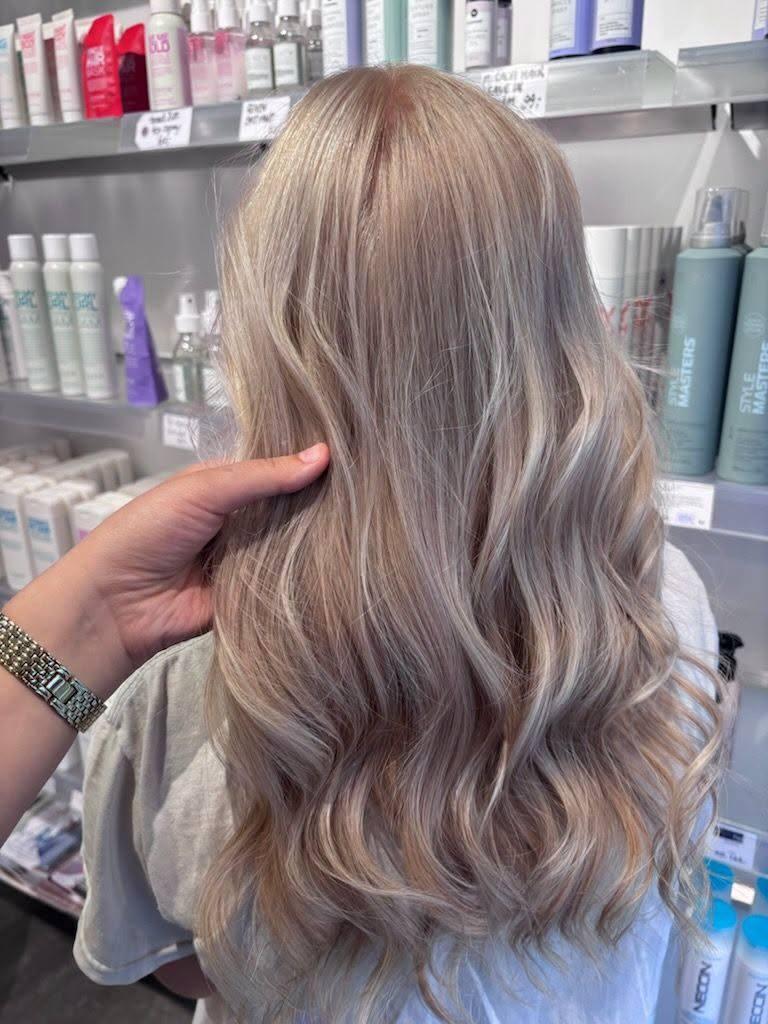 En person med langt, bølget hår i lys blond farve, mens de står foran hylder med hårprodukter.