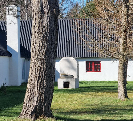 En hvid bygning med røde vinduer og en grill i haven, omkranset af store træer.