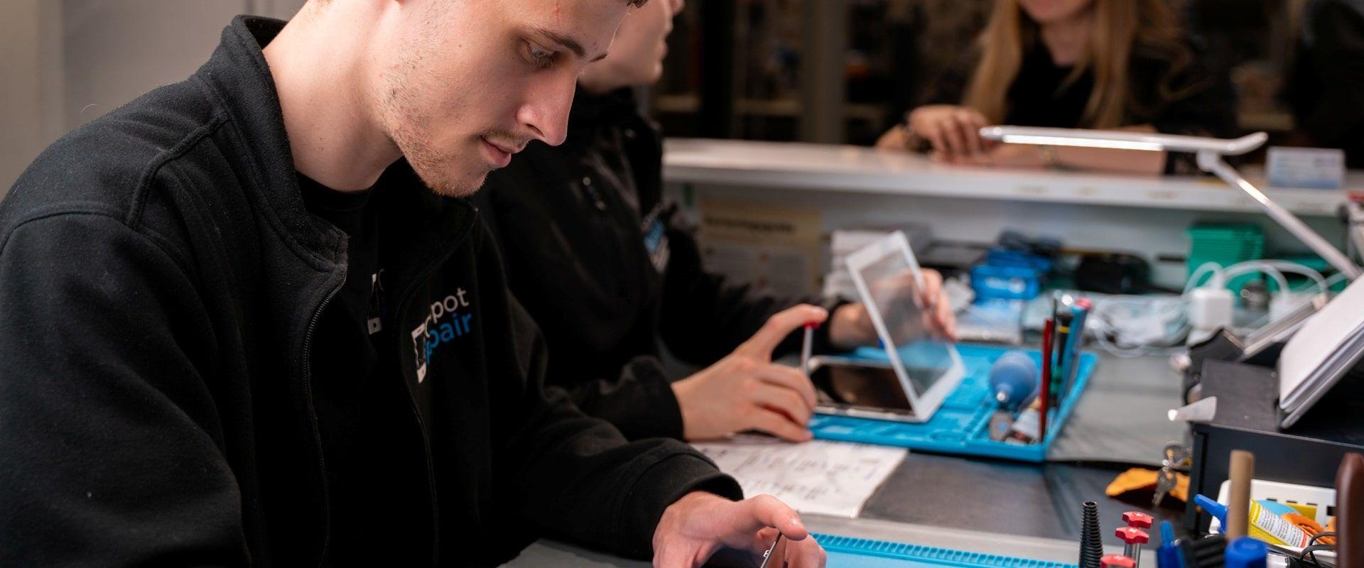 En ung mand arbejder koncentreret med elektronik i et reparationsværksted, mens en kvinde bruger en tablet i baggrunden.