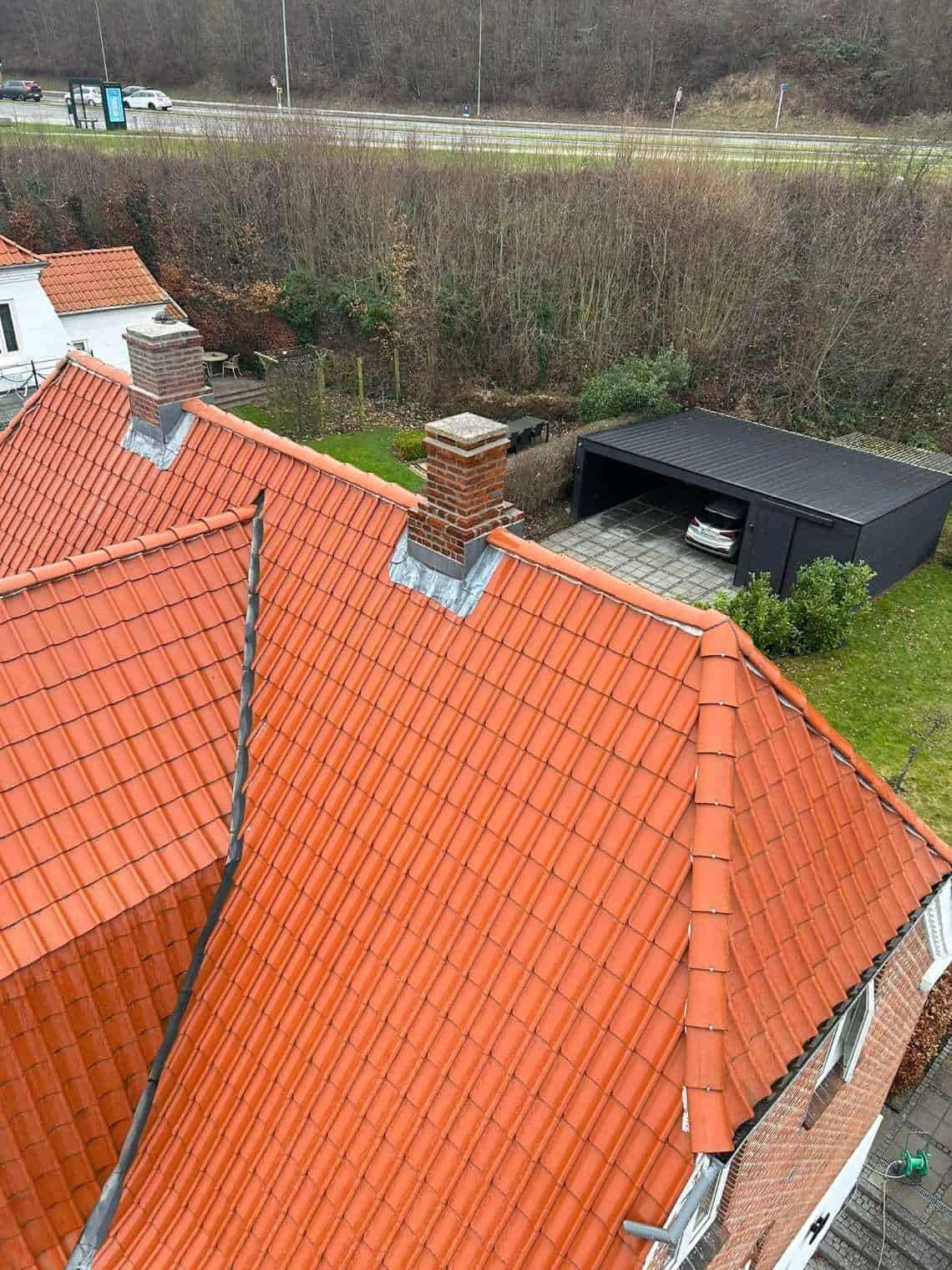 Tag med røde teglsten og skorsten, set fra oven, med en garage i baggrunden og skov i nærheden.