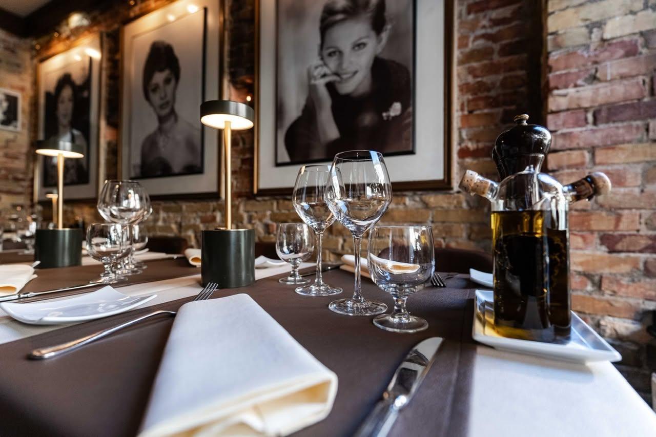 Et elegant restaurantbord med glas, bestik og olivenolie, omgivet af store portrætter af kvinder på væggene.