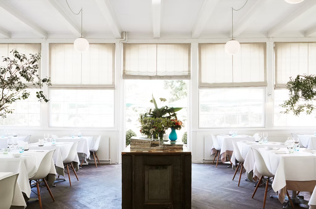 Lyst restaurantlokale med hvide borde, planter og en blomsterdekoration på et centralt bord.