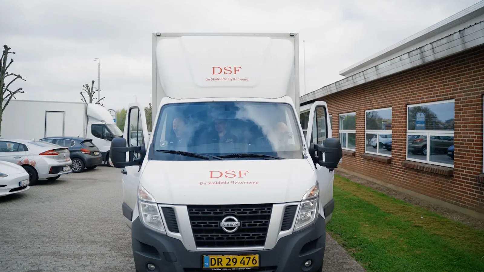 En hvid varevogn med DSF-logo parkeret foran en bygning med vinduer.