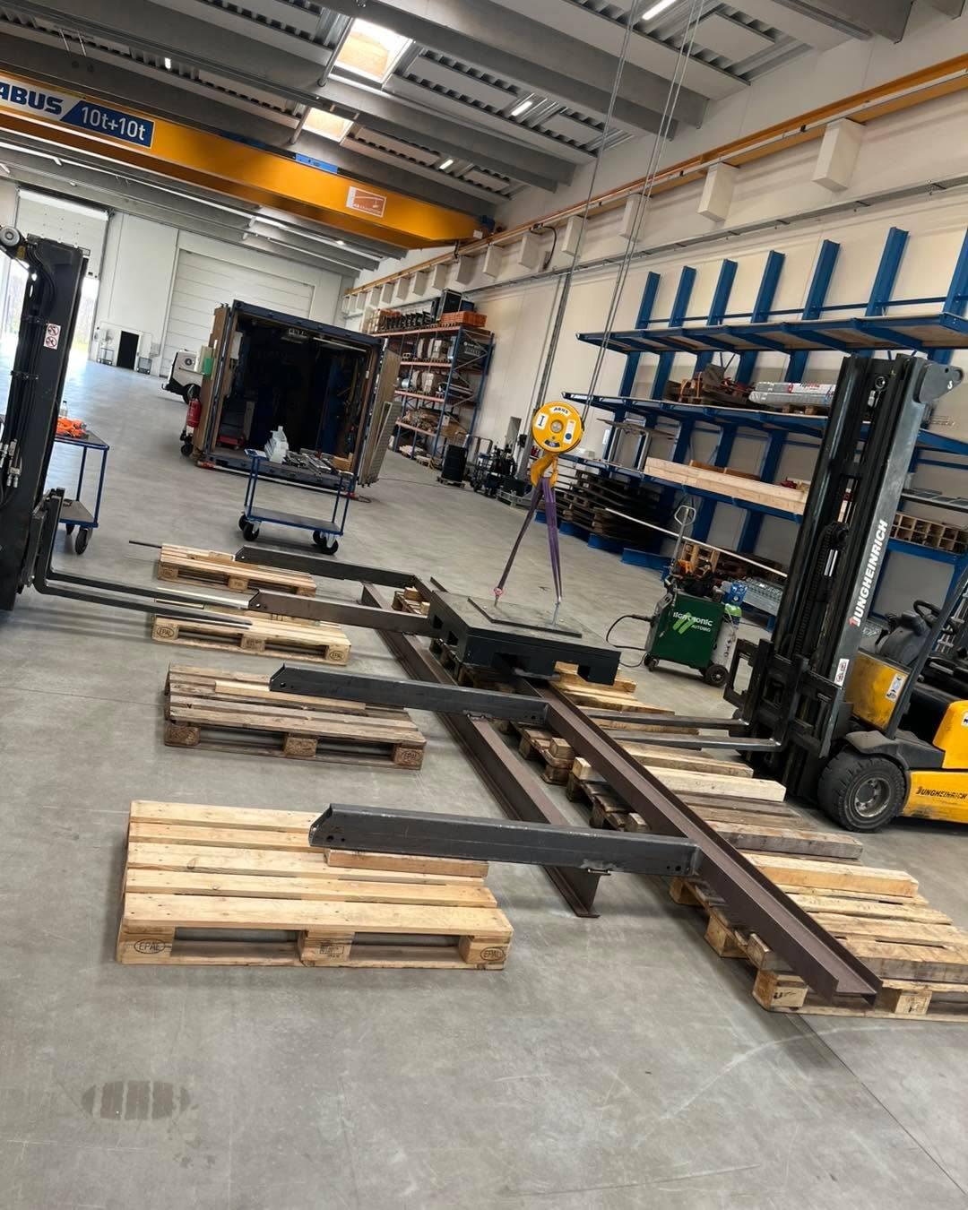 Lagerområde med palletrucks og metalramme på træpaller i en stor hal med hylder i baggrunden.