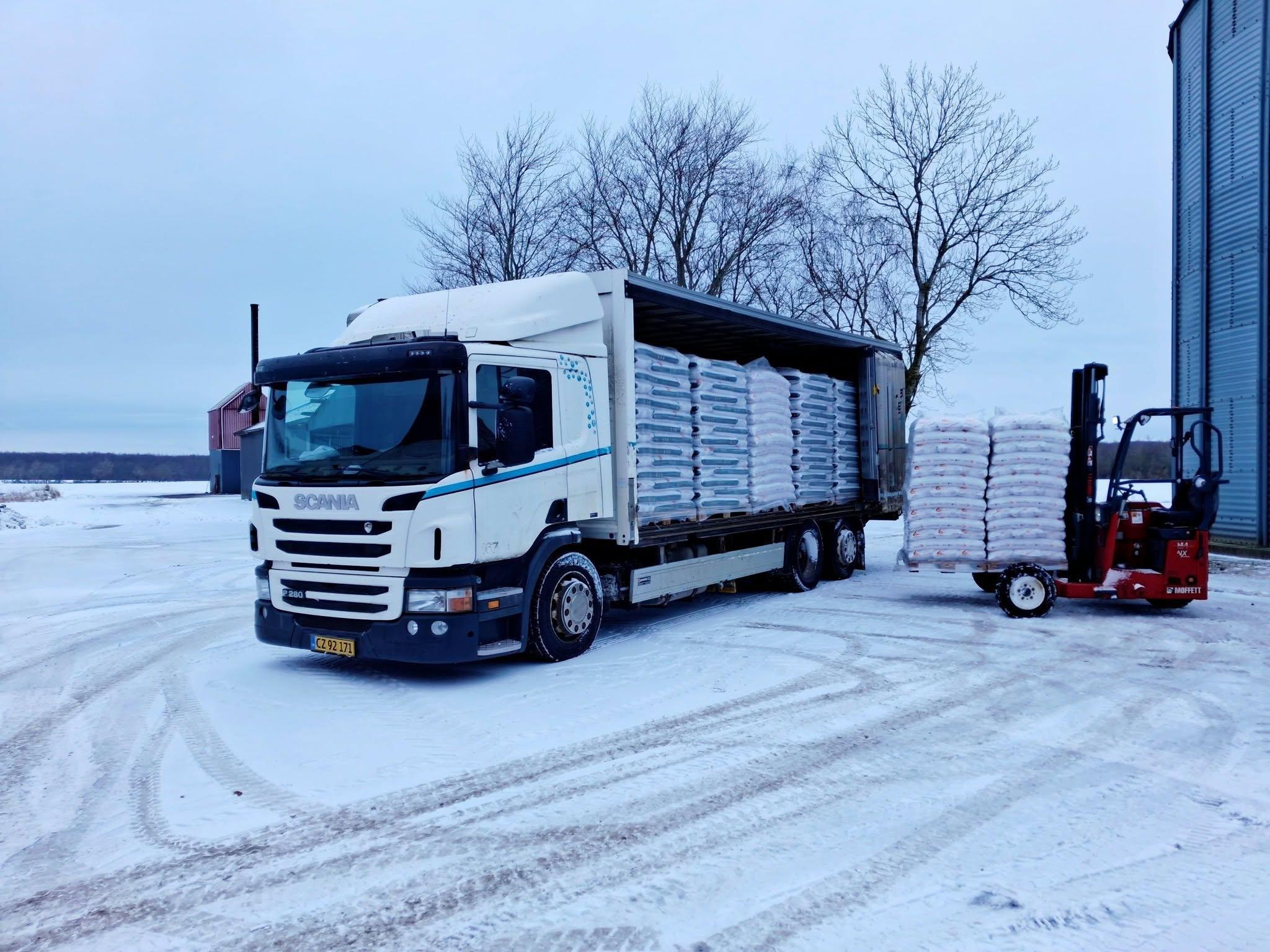 Lastbil med trailer fyldt med kasser på en sneklædt gård, mens en gaffeltruck står klar til at laste.