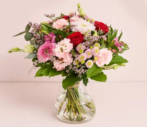 En farverig buket blomster i en vase med vand, bestående af roser, gerberaer og krysantemum.