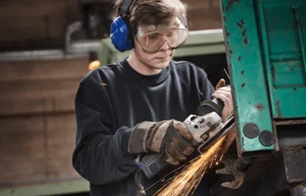 En person med beskyttelsesbriller og høreværn sliber metal med en vinkelsliber, gnister flyver.