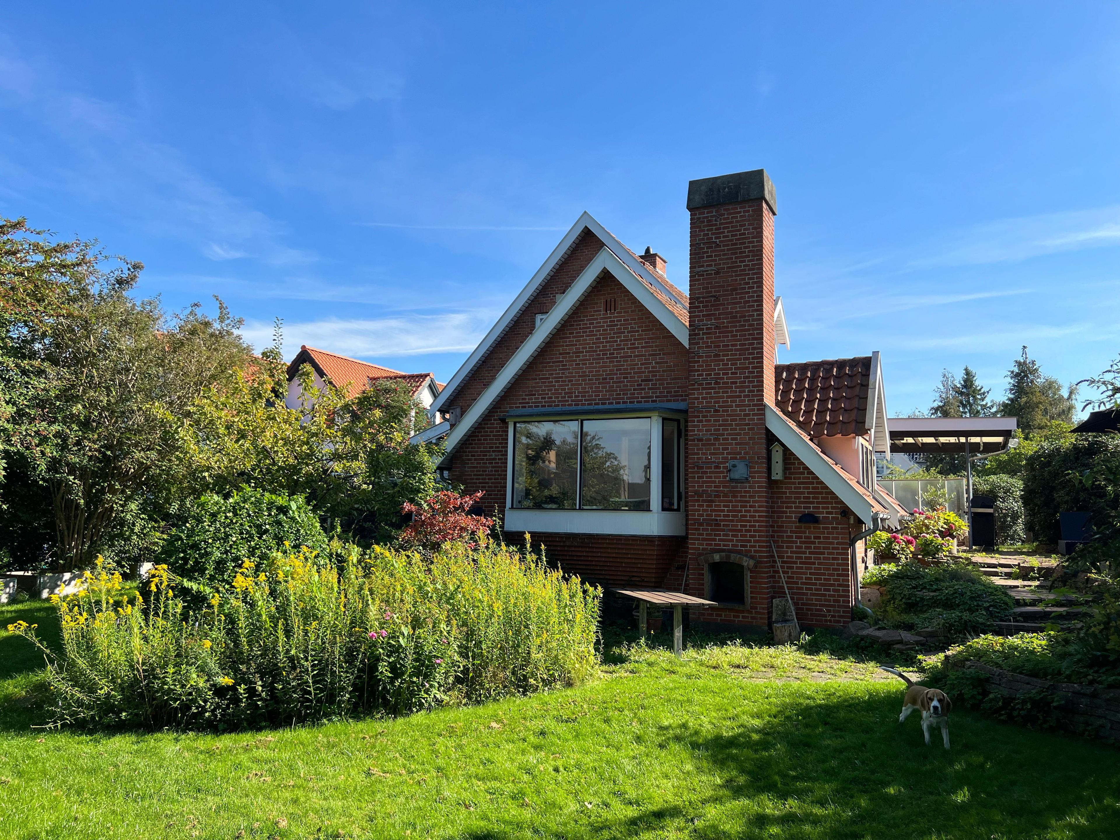 Et rødt hus med en have fyldt med blomster og buske under en blå himmel.