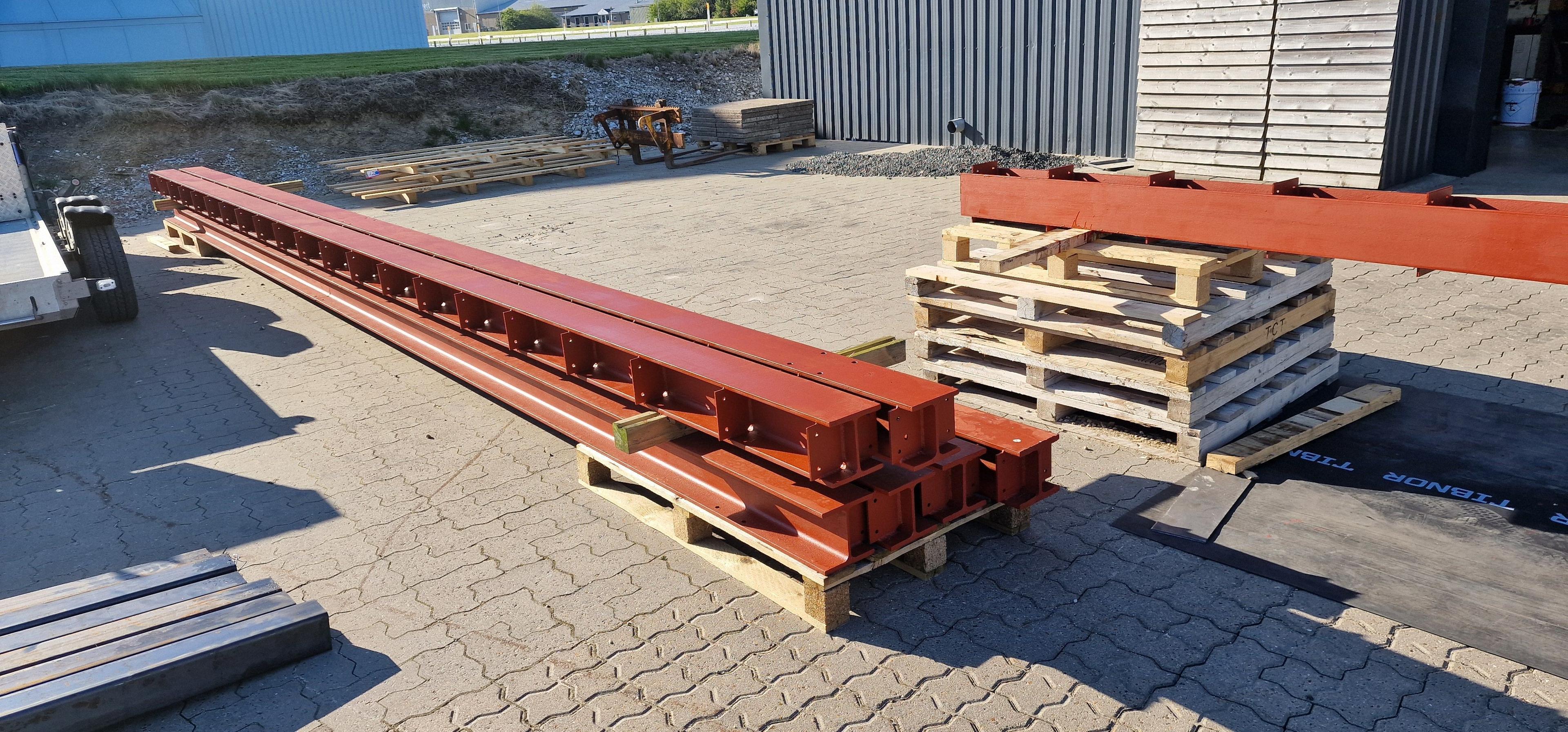 Lange stålbjælker stablet på paller ved en bygning på en byggeplads.