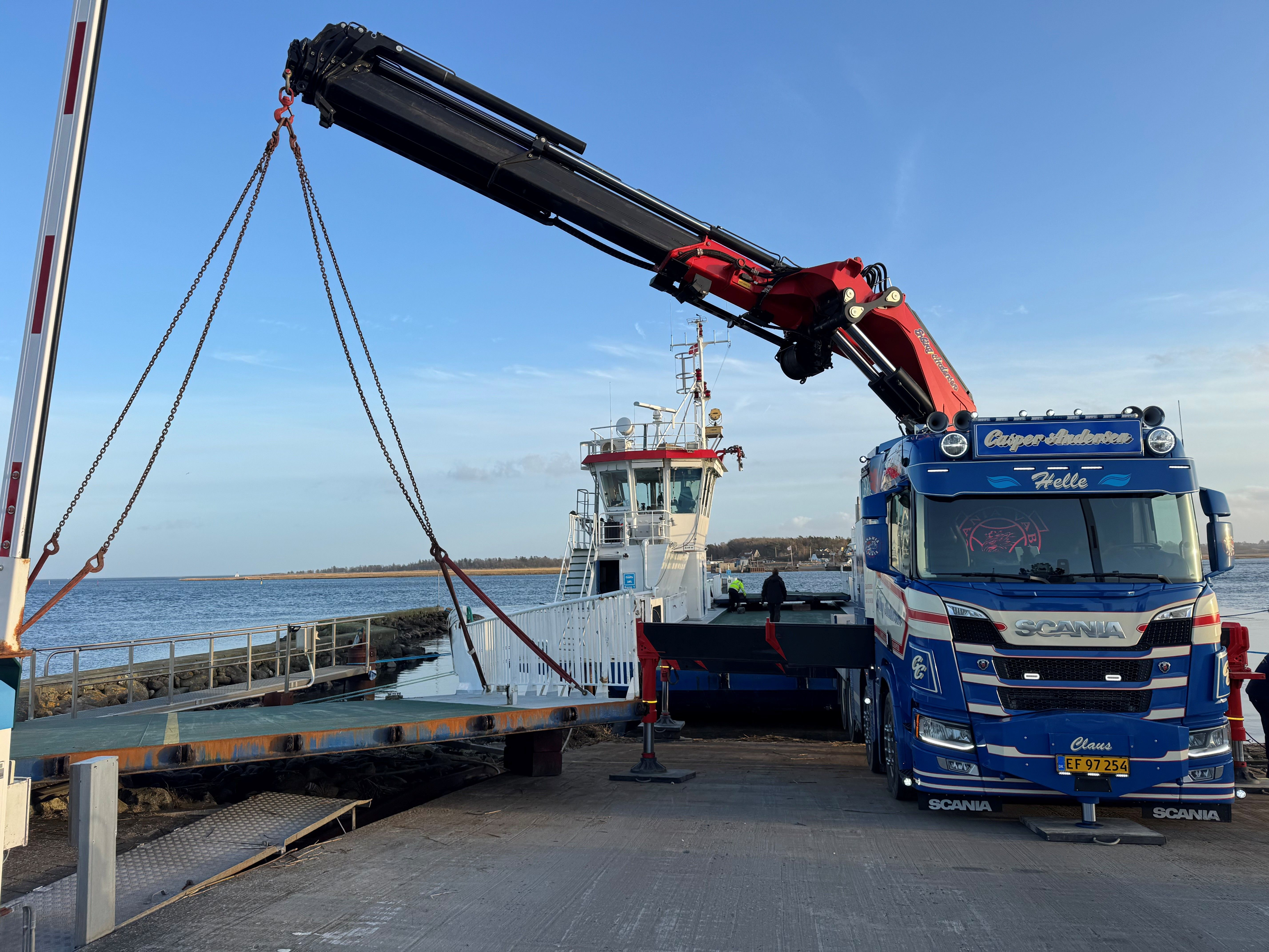 En lastbil med kran løfter en båd ved en havn med vand og klipper i baggrunden.