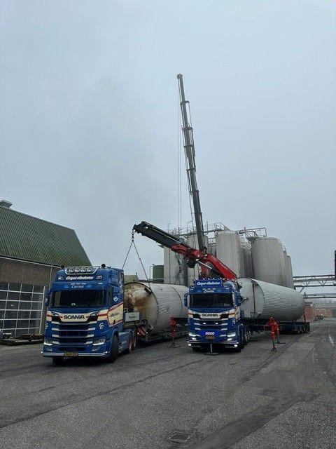 To lastbiler med kraner løfter store tanke på en industriel plads under en grå himmel.