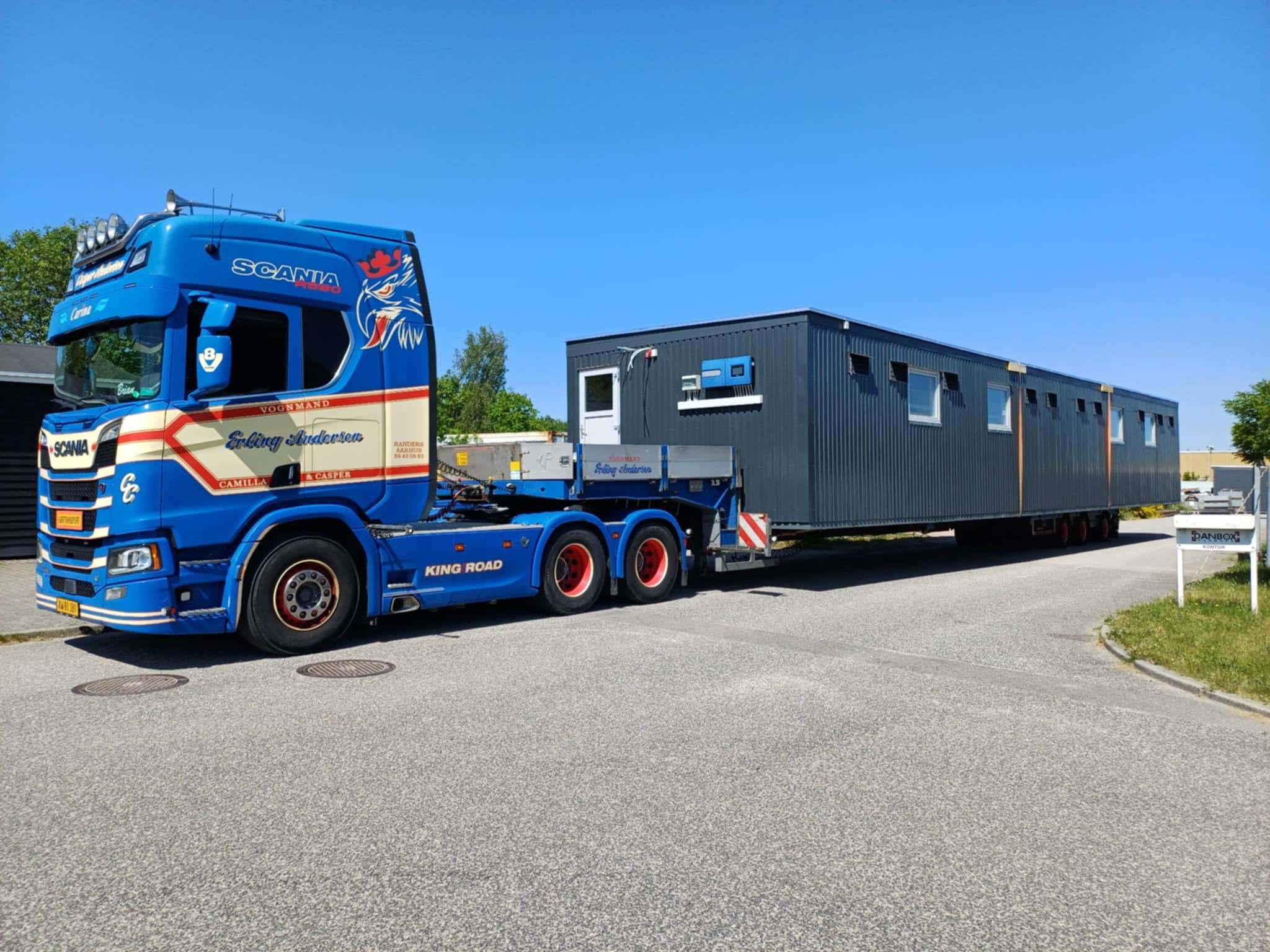 En stor lastbil trækker en lang container med vinduer på en solrig vej.