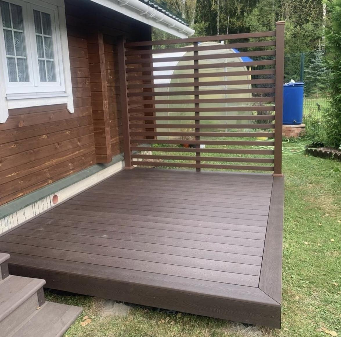 Terrasse i træ med en moderne skillevæg, placeret ved siden af et hus i en grøn have.
