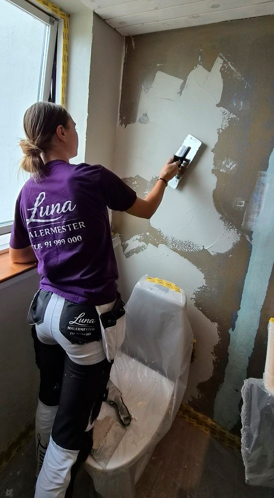 En kvinde maler en væg i et badeværelse, iført en lilla t-shirt med teksten "Luna" og malerudstyr.