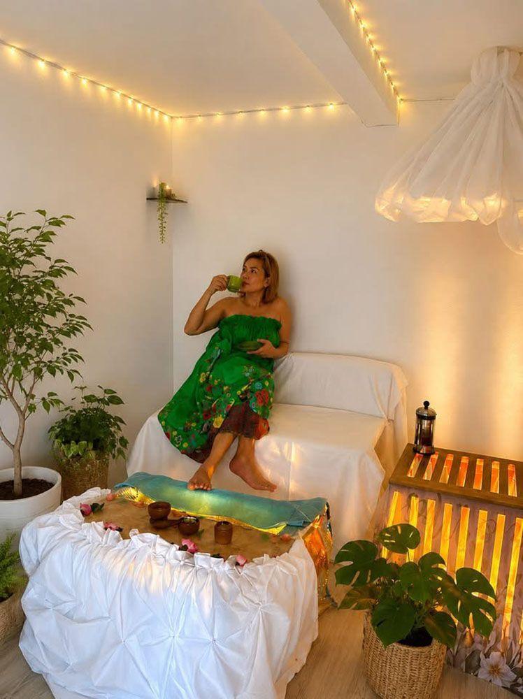 En kvinde i en grøn kjole sidder på en sofa i et lyst rum med planter og dæmpet belysning.