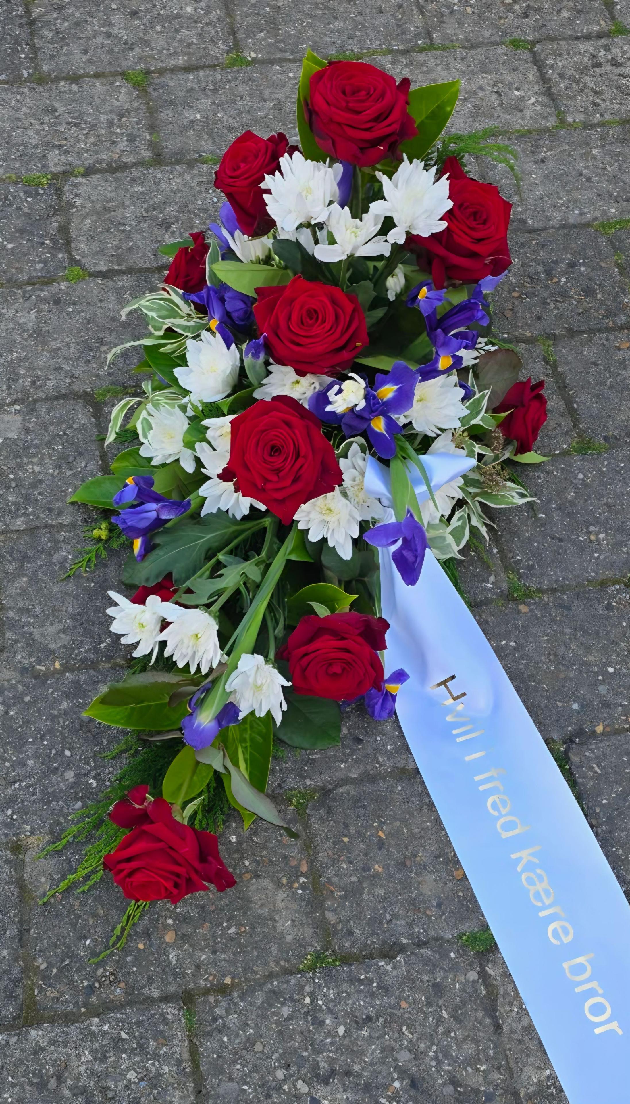 En smuk krans med røde roser, hvide blomster og blå iris, prydet med et blåt bånd.