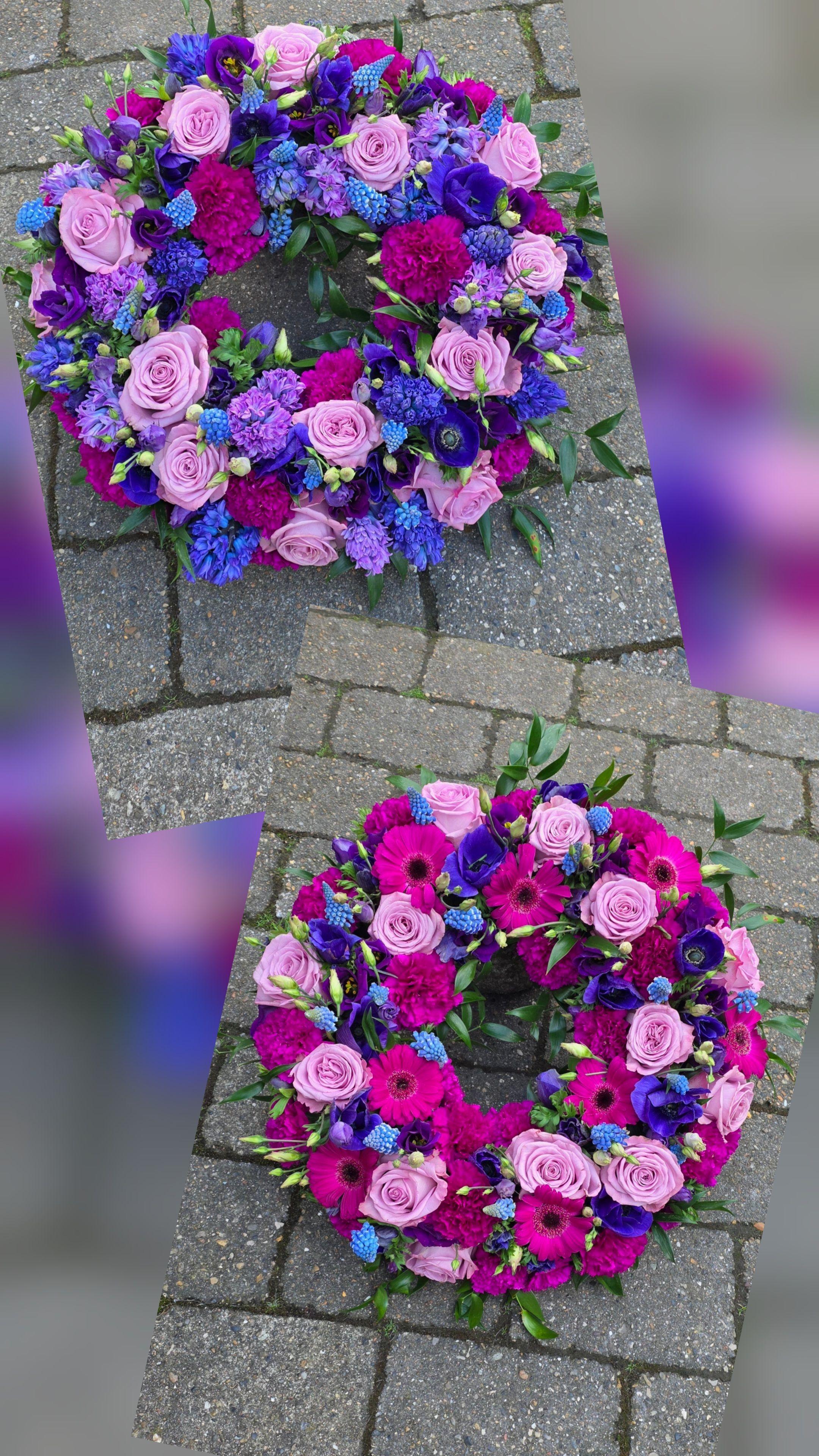 Farverig krans med lyserøde roser, lilla anemoner og blå blomster på en grå flisebelægning.