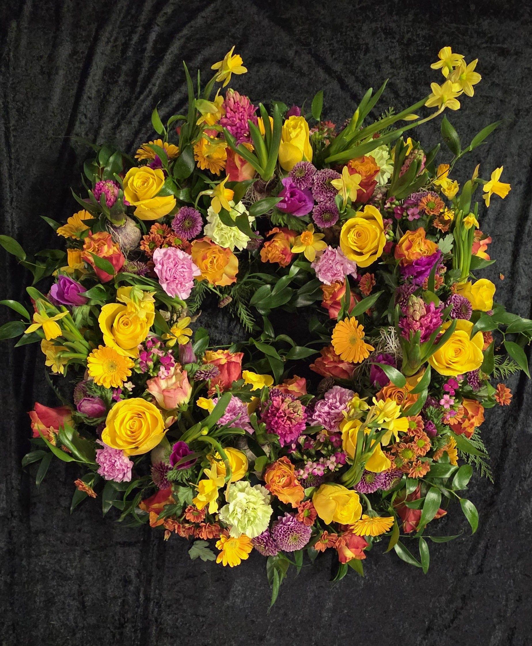 En farverig blomsterkrans med gule roser, lilla nelliker og orange blomster på en sort baggrund.