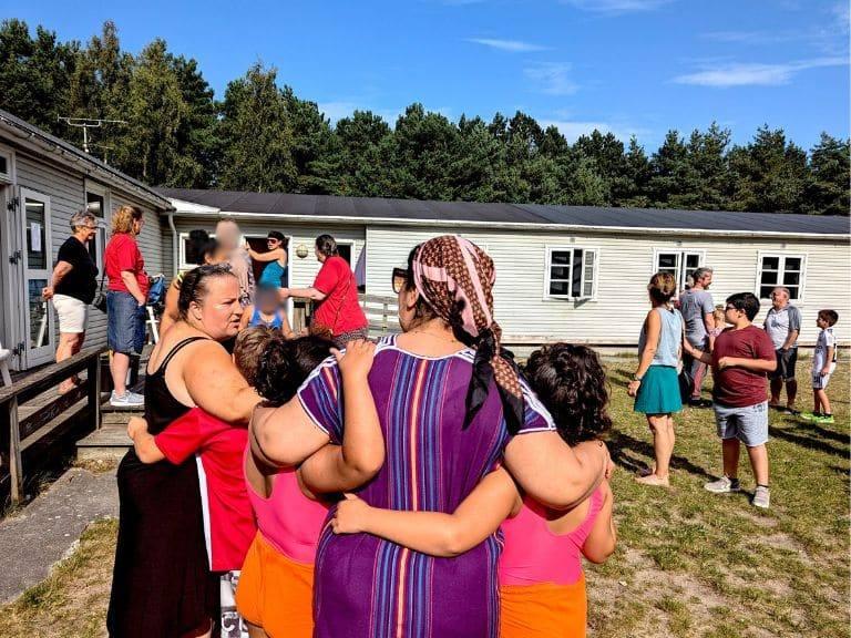 Gruppe af mennesker, der står sammen i en gård, med børn og voksne, der interagerer i solrigt vejr.