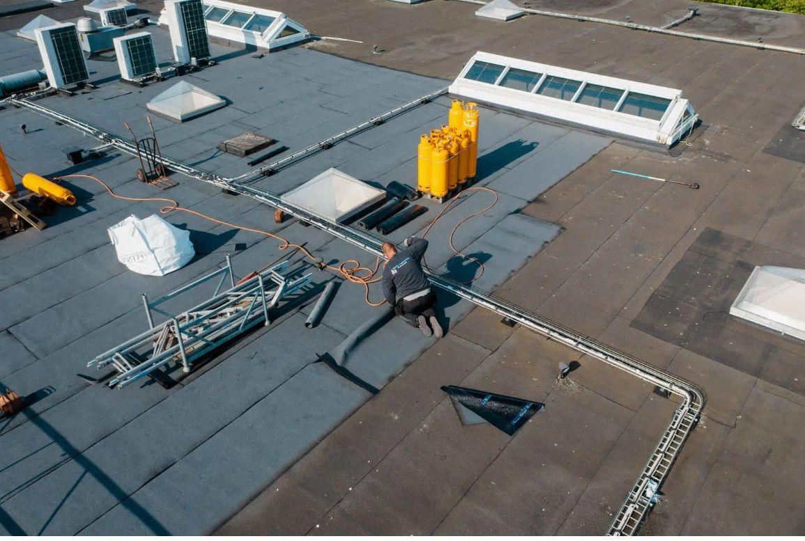 En håndværker arbejder på et fladt tag med solpaneler og ventilationsanlæg i baggrunden.
