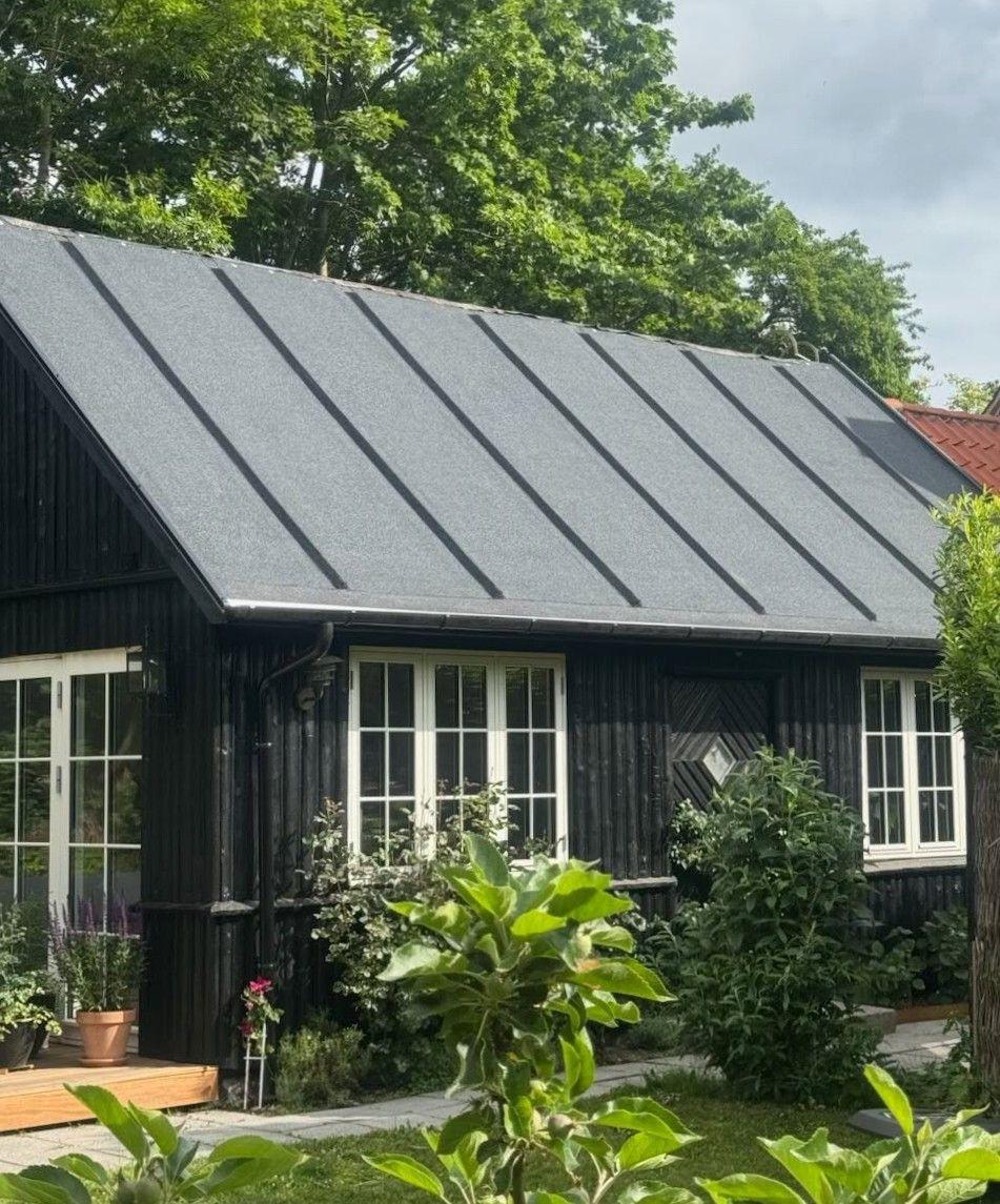 Sort træhus med skrå tag og store vinduer omgivet af grønne planter og træer.