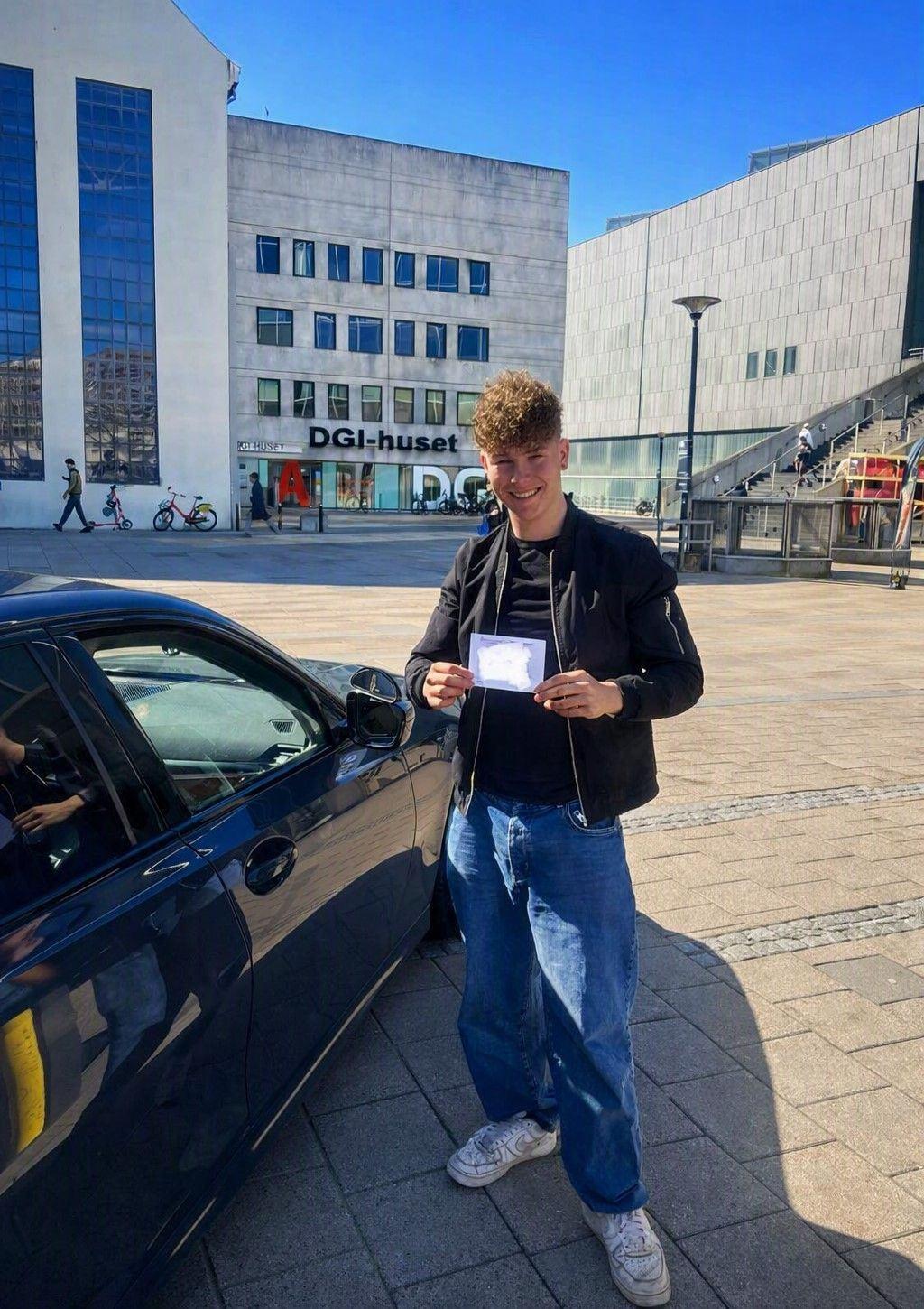 Ung mand står ved en bil og holder et dokument op, med moderne bygninger i baggrunden.
