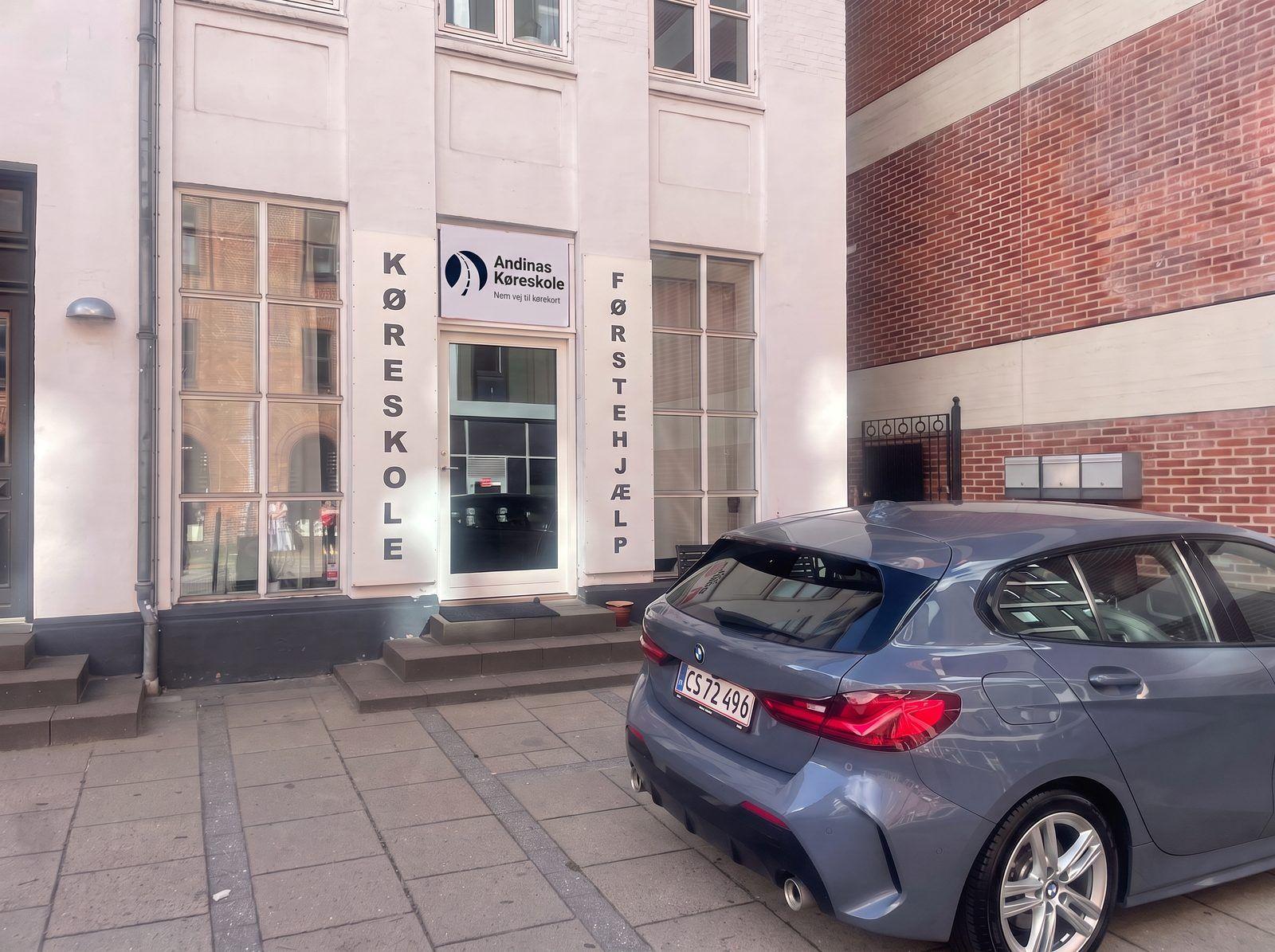 Køreskole facade med skilte og en grå bil parkeret foran bygningen.