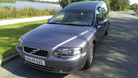 En grå Volvo ligvogn parkeret ved en vej med grønne træer og en sø i baggrunden.