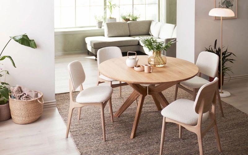 Et rundt træbord med fem stole i en lys stue med planter og en sofa i baggrunden.