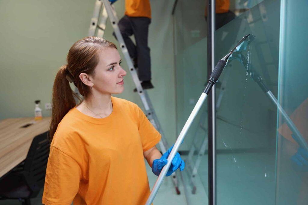 En kvinde i orange t-shirt vasker vinduer med en lang stang, mens en anden person står på en stige i baggrunden.