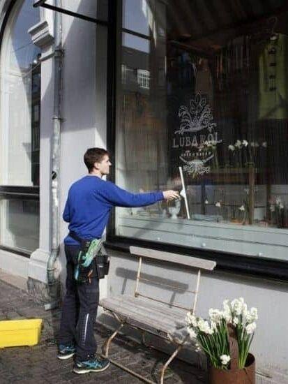 En mand vasker vinduet på en butik, mens han står foran en bænk og blomster i en kasse.