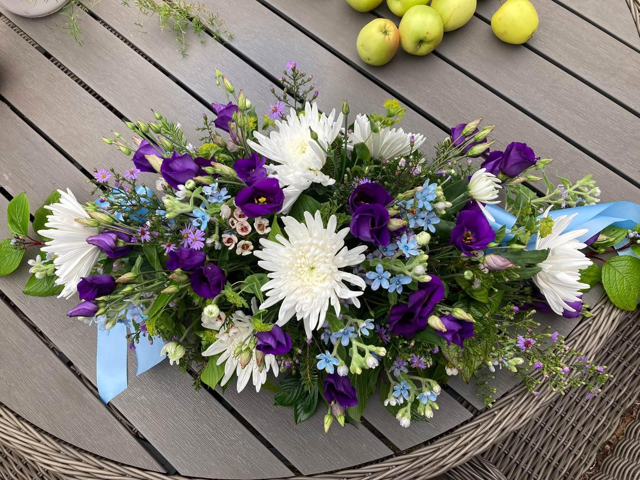 En farverig blomsterdekoration med lilla, hvide og blå blomster, prydet med bånd, ligger på et træbord.