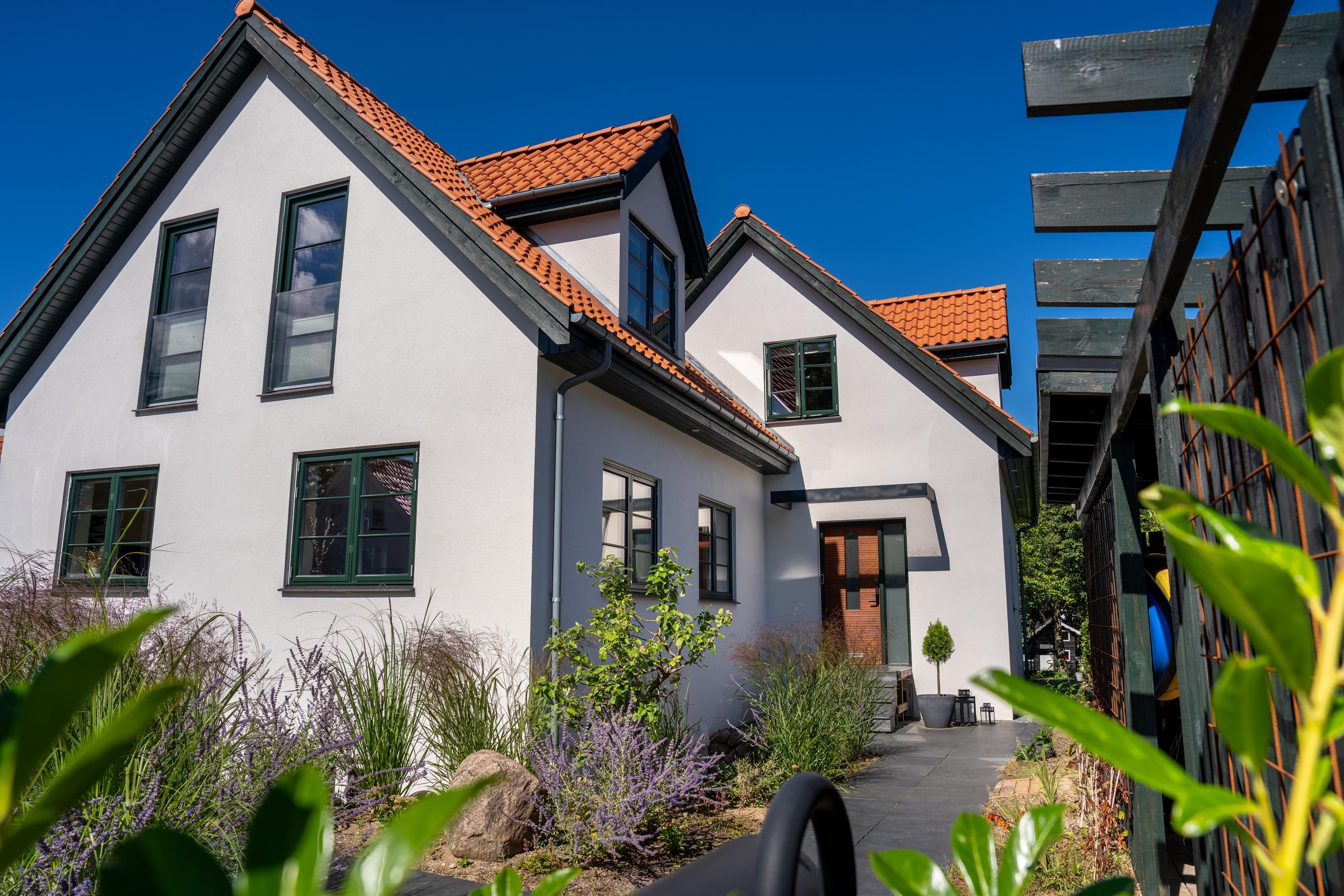 Moderne hvidt hus med rødt tag, grønne vinduer og velplejet have under en klar blå himmel.