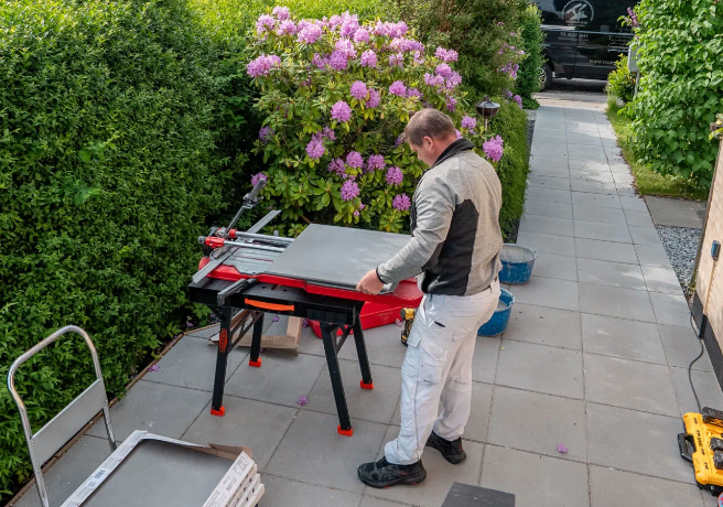 En mand arbejder med en arbejdsbænk på en flisebelagt gårdhave omgivet af blomstrende rhododendron.