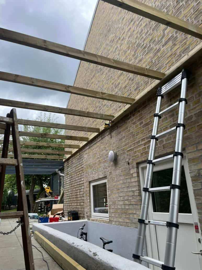 Byggeri af en terrasse med træbjælker og en stige ved en murstensvæg.