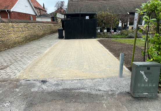 Ny belægning på indkørslen med grus og en sort garage i baggrunden, omgivet af hække og planter.