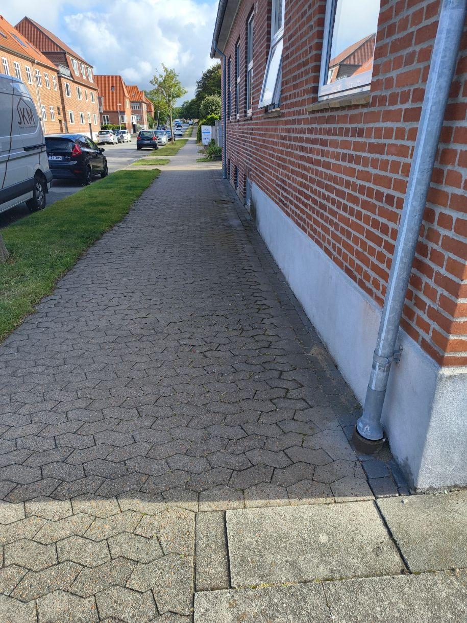 En fortov langs en murstensbygning med græsplæne og parkerede biler i baggrunden.