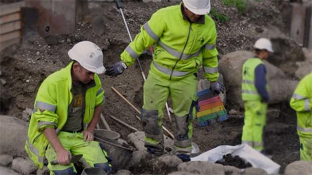 Arbejdsfolk i neonfarvet arbejdstøj arbejder på en byggeplads med grus og sten.