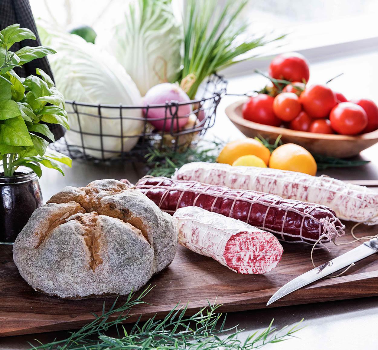 Friskbagt brød, pølser, tomater, citrusfrugter og grøntsager på et skærebræt i et lyst køkken.