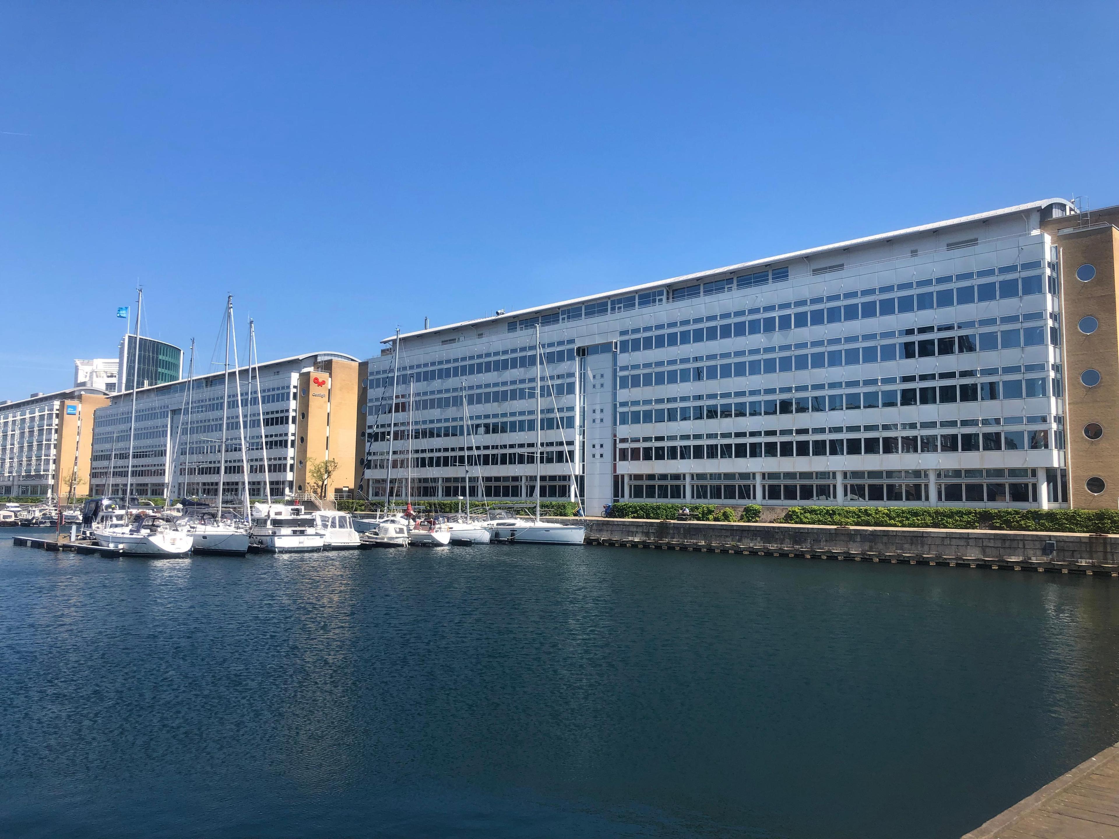 Moderne bygninger ved havnen med sejlbåde fortøjet i vandet under en klar blå himmel.