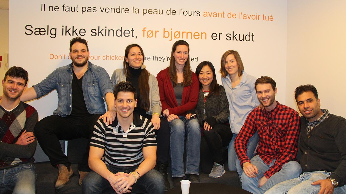 Gruppe af unge mennesker smilende foran en væg med tekst på dansk og fransk, der opfordrer til at tænke før man handler.