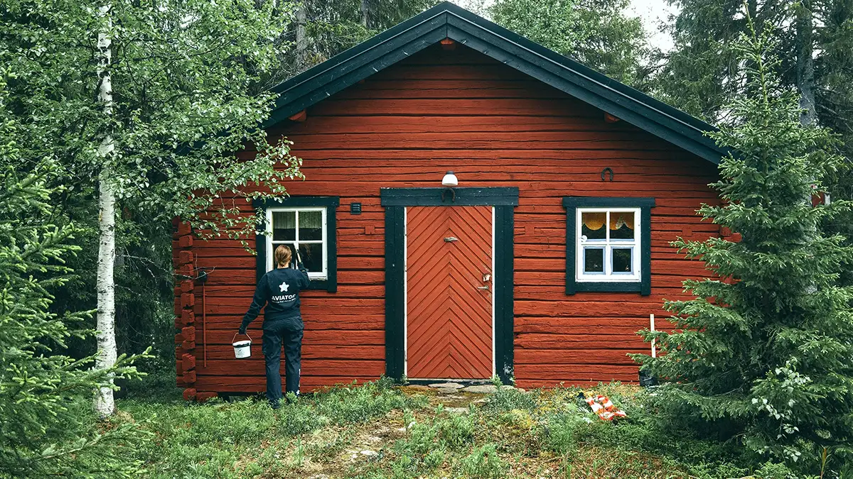 En person der holder en spand står foran et rødt hus omgivet af græs og træer.
